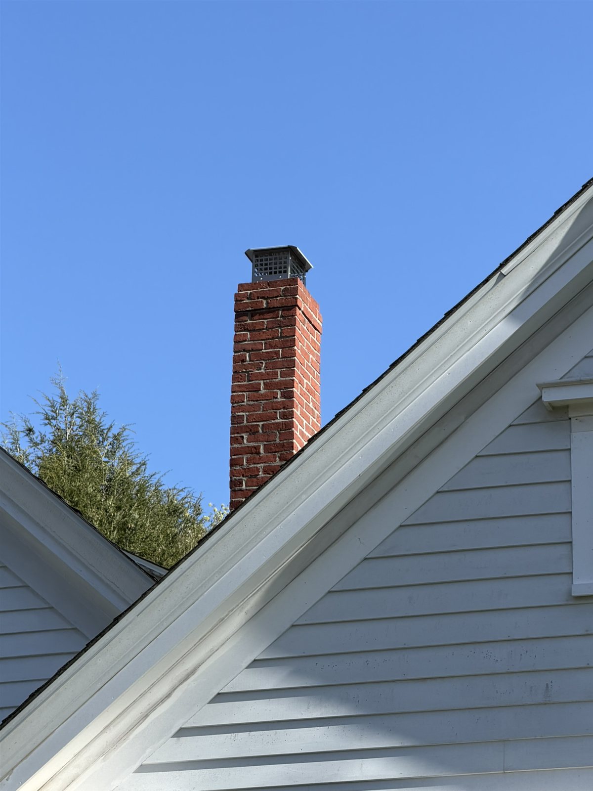Stainless steel mesh chimney cap installed on a tall brick flue against clear blue sky.